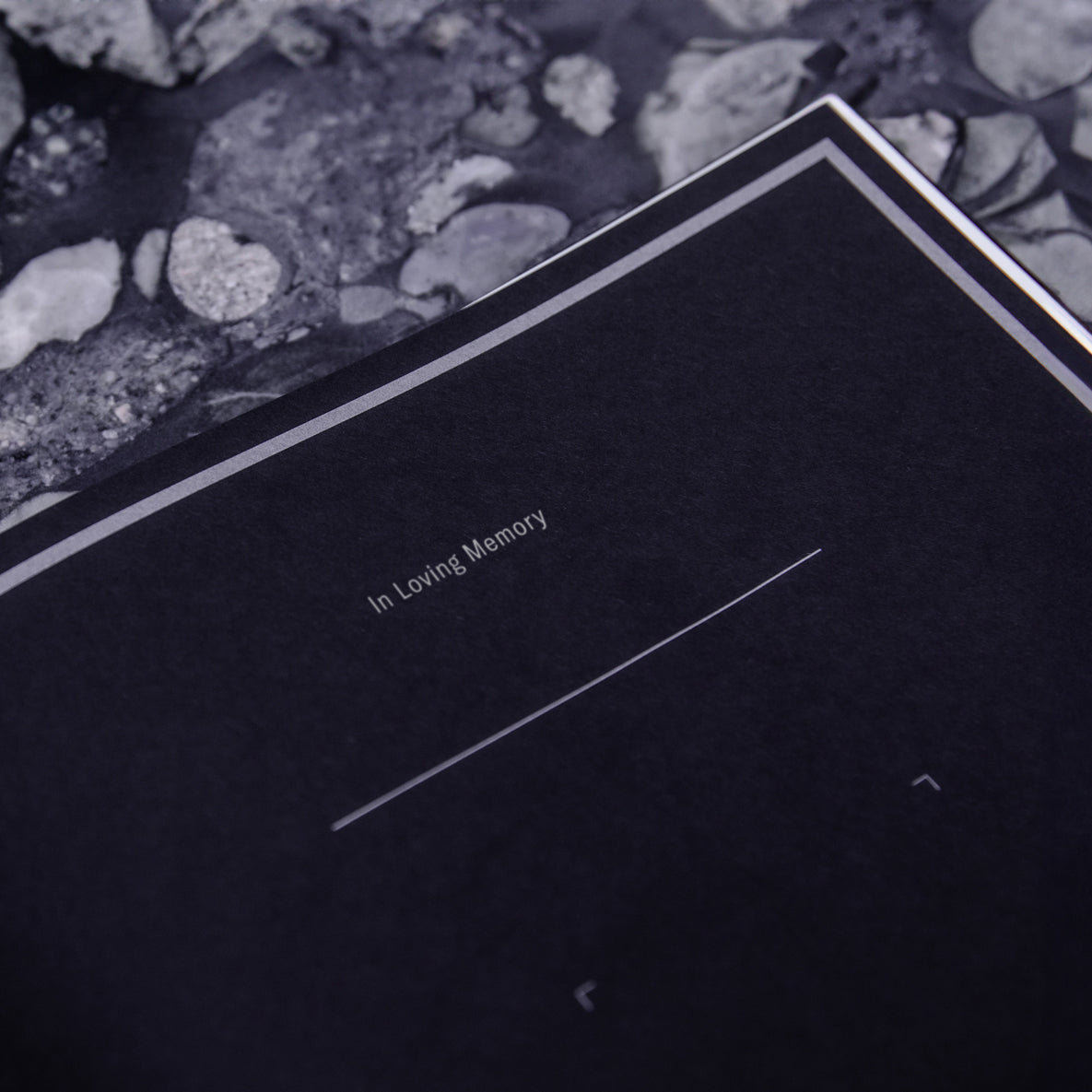 A close-up of a black memorial keepsake box or book cover with the words 'In Loving Memory' and a blank line for personalization, resting on a dark stone background.