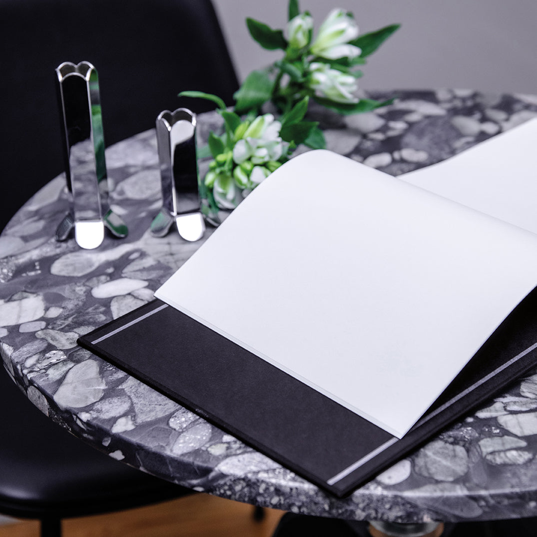 A black hardcover notebook or binder with a large blank white page, styled on a black-and-white marble table next to two mirrored rectangular stands and a small green plant.