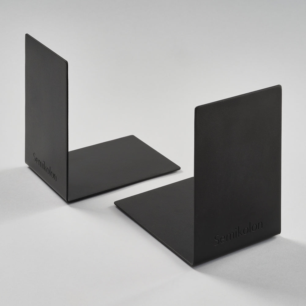 Two matte black metal bookends on a light gray studio background, each with a rectangular backplate and flat base. The brand name 'Semikolon' is subtly embossed on the lower right of each plate.