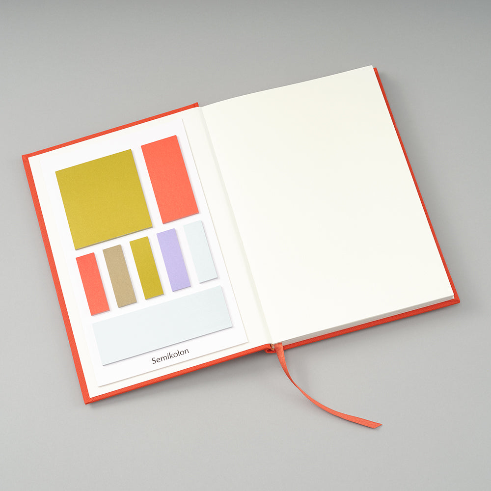 An open hardcover notebook with a coral-orange cover and ribbon bookmark, showing a printed color card with geometric color blocks on the left page and blank white paper on the right, photographed on a light gray background.