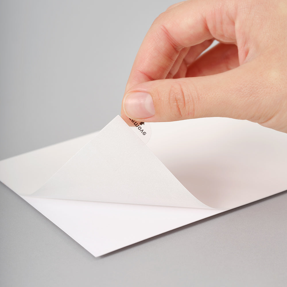 A close-up of a hand peeling back the top flap of a white square sheet, showing a small printed brand mark on the underside against a neutral gray background.
