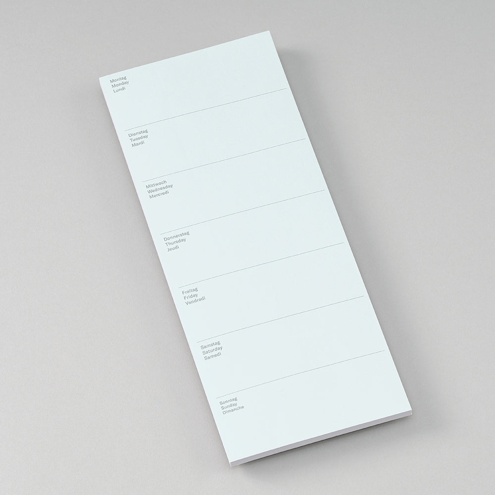 A white daily planner notepad with columns for Monday through Sunday and blank lines for notes, shown on a neutral light gray background.